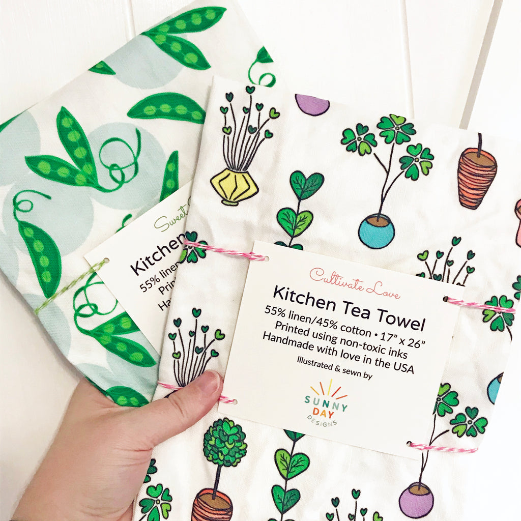 2 colorfully printed and fun tea towels are shown packaged and held in a hand in front of a white background. 1 tea towel is printed with green sweet peas. 2nd dish towel shows colorful potted plants with heart shaped leaves.