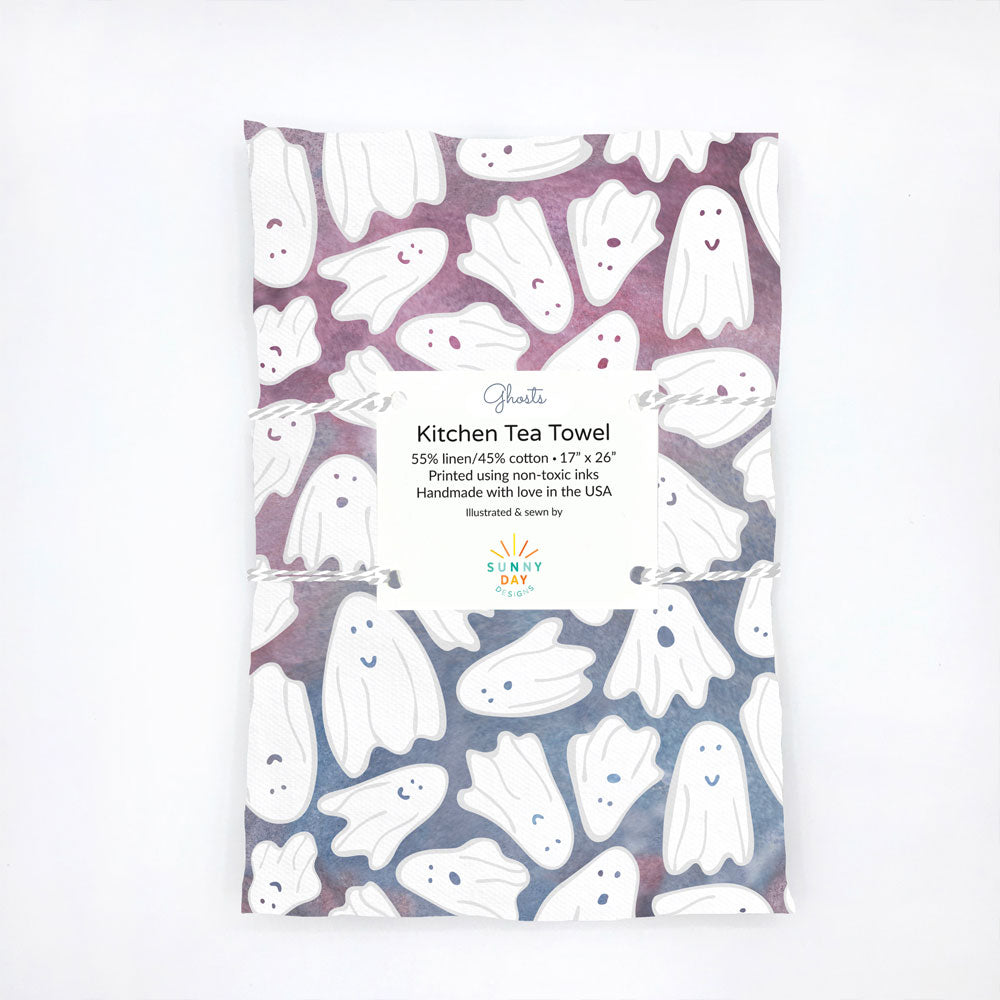 A handmade, fun kitchen tea towel featuring a variety of happy Ghosts floating on a blue and purple watercolor background. This Halloween-themed, linen/cotton dish towel is made in the USA by Sunny Day Designs