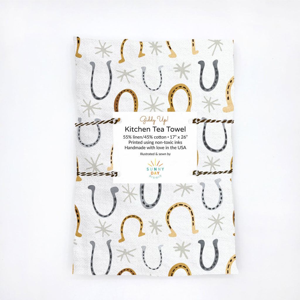 A packaged, handmade linen/cotton tea towel printed with a gray and gold watercolor horseshoe print by Sunny Day Designs.