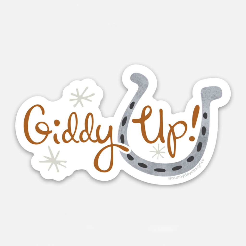 a horse riding-themed die cut equestrian vinyl sticker is shown on a blank white background with the words "Giddy Up!" in brown hand lettered text and a gray watercolor horseshoe design.