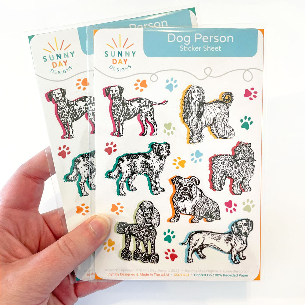 A hand holding 2 packaged "Dog Person" sticker sheets by Sunny Day Designs, which feature 7 different dog breeds and numerous colorful paw print stickers. Printed in the USA on 100% recycled uncoated paper & designed by Sunny Day Designs.