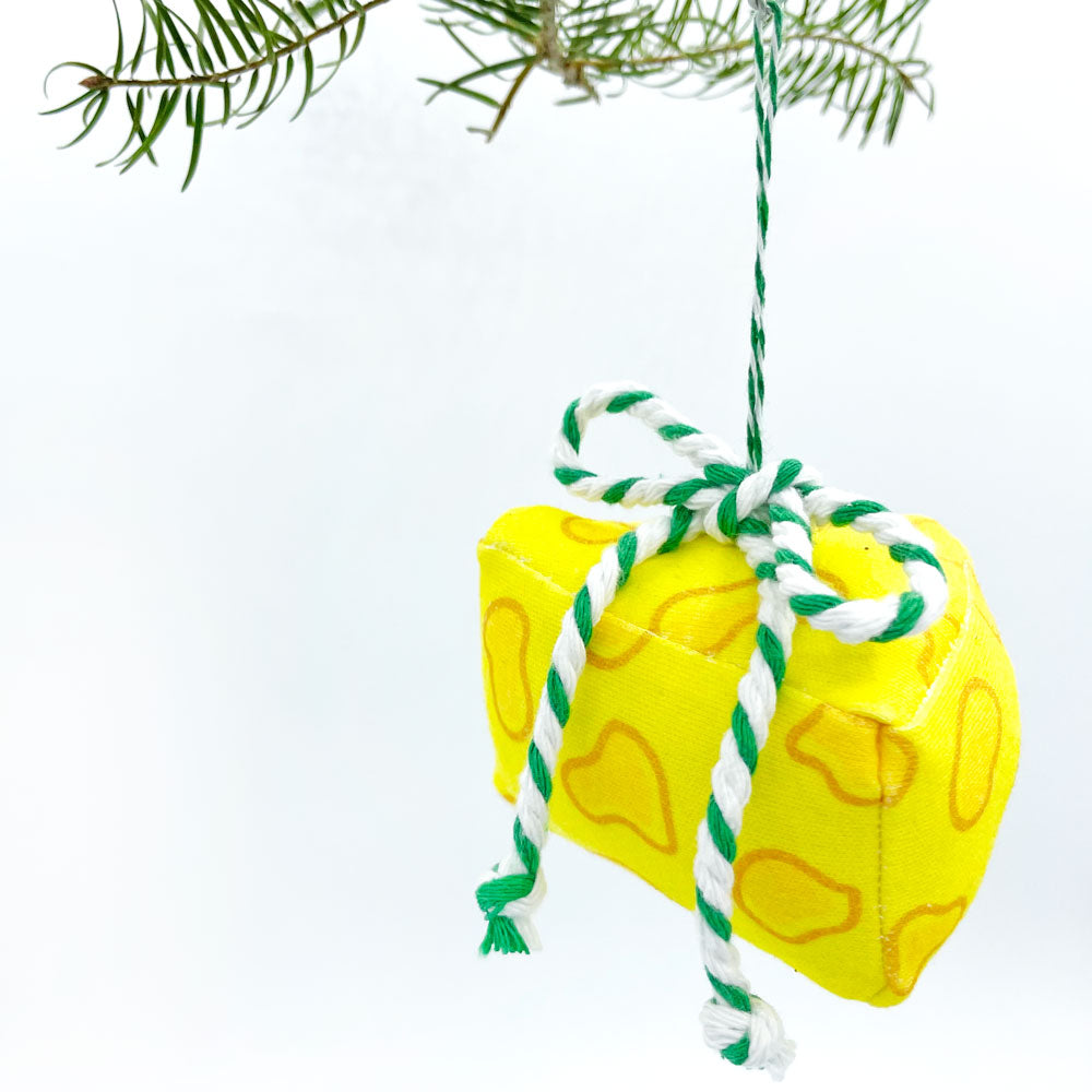 3D handmade yellow Cheese Christmas Ornament is made from 100% organic cotton fabric and features a green and white striped baker's Twine Bow at the top. This holiday ornament is made in the USA by Sunny Day Designs and the image shows it hanging from a Christmas tree branch against a white background.
