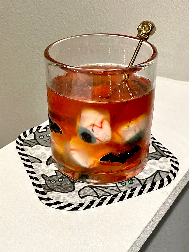 Spooky Halloween Cocktail in Bat Old Fashioned Glass with Skull Drink Stirrer, Eyeball ice cubes, and cute handmade Benevolent Bats printed fabric cocktail napkin by Sunny Day Designs.