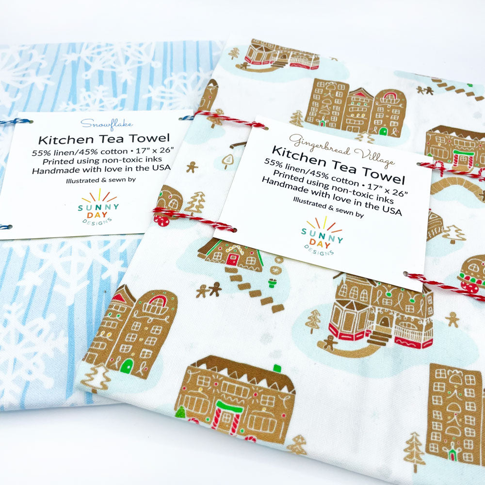 This fun set of 2 handmade holiday tea towels by Sunny Day Designs features 1 blue/white Snowflake winter printed dish towel & 1 whimsical Gingerbread Village printed kitchen towel. Made in the USA & shown at an angle while folded & packaged on a white background.