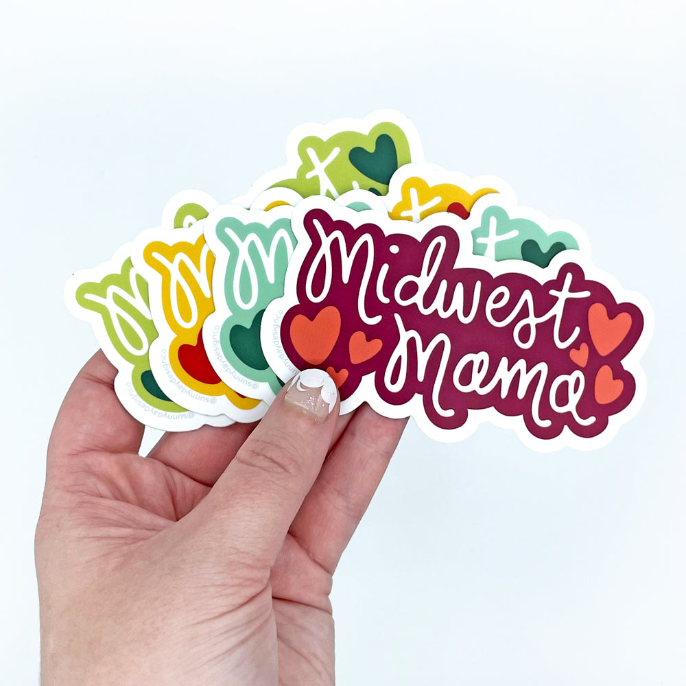 Midwest Mama Vinyl Stickers in 4 Color Options. These fun stickers are shown held in a hand in front of a white background. Designed and made in the USA by Sunny Day Designs