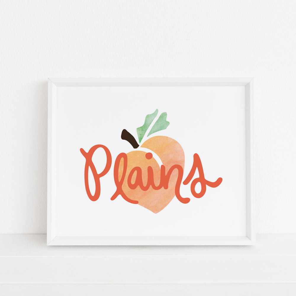Plains Peach Georgia-themed 8x10 art print is framed ina white picture frame againsta a white background. Plains Peach artwork design features an orange, green, and brown watercolor peach design with dark orange handlettered "Plains" city name on top of peach. Professionally printed on watercolor paper. Made in the USA by Sunny Day Designs