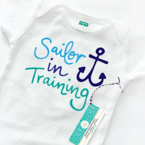 Sailor In Training organic cotton baby onesie by Sunny Day Designs features an anchor and hand lettered "Sailor In Training" text in shades of light blue, navy blue, and turquoise. Organic cotton. made in the USA.