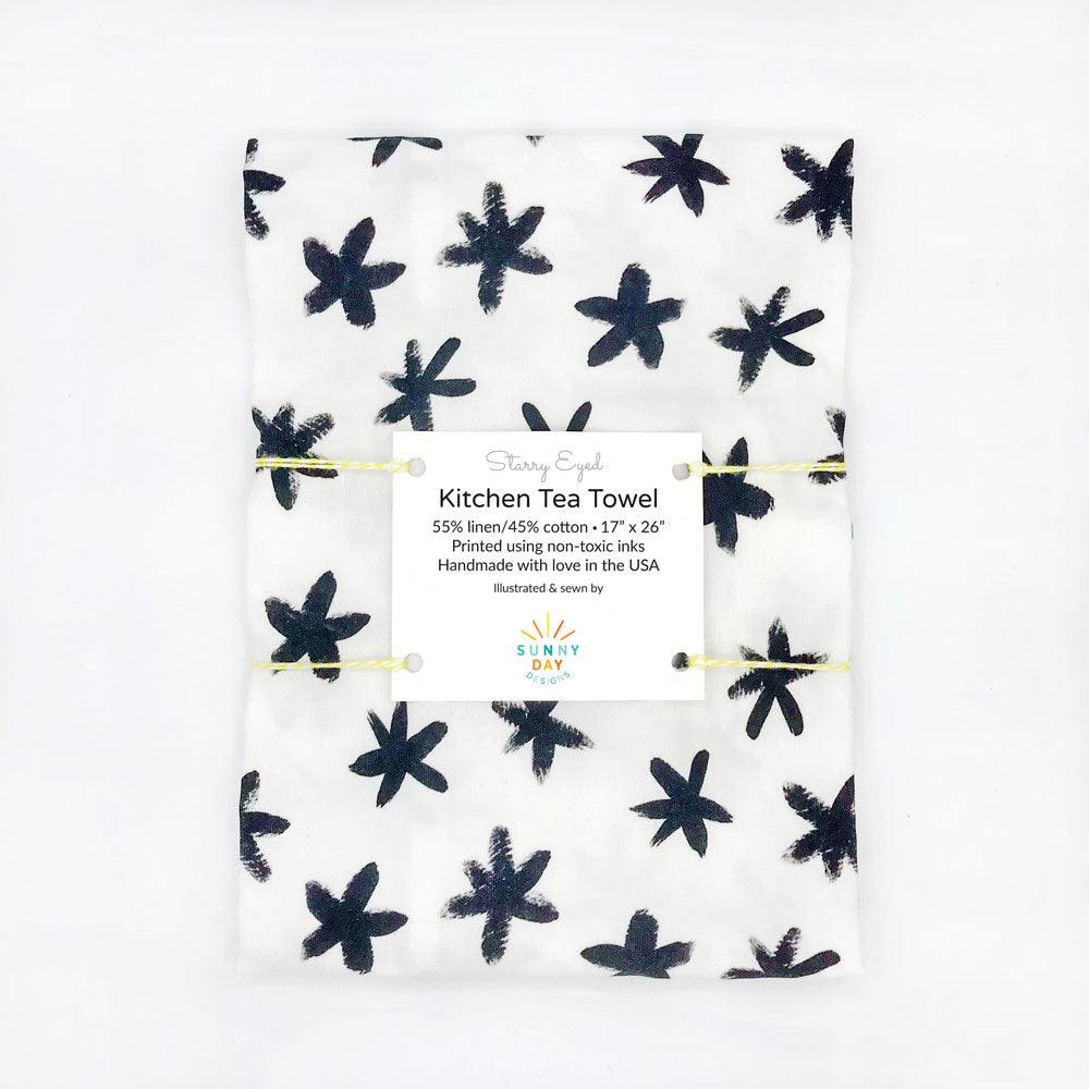 Our black and white Starry Eyed printed kitchen towel includes black brushstroke stars on a white fabric. This packaged dish towel by Sunny Day Designs is made in the USA and photographed on a white background.