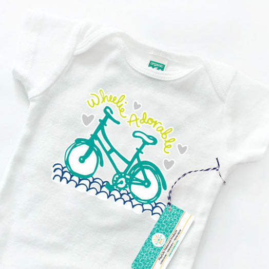 Close up image of a bicycle themed organic cotton baby onesie by Sunny Day Designs. This Wheelie Adorable baby bodysuit makes a perfect baby shower gift for cyclist parents and bike enthusiasts. Teal bicycle on blue street with lime green hand lettered text and gray hearts. made in the USA in Madison Wisconsin.