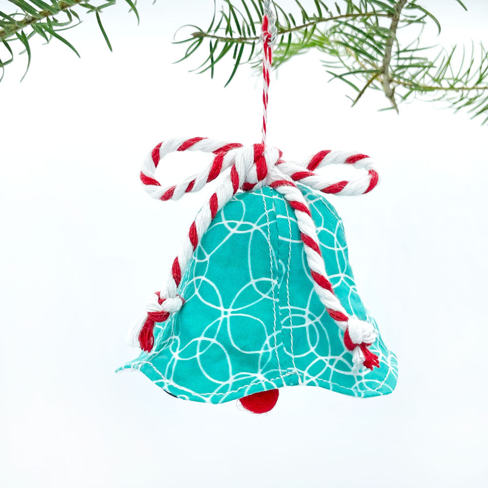 A turquoise, red, and white holiday bell shaped Christmas ornament (made in the USA from stitched fabric by Sunny Day Designs) is hanging from a green Christmas tree branch against a white background.