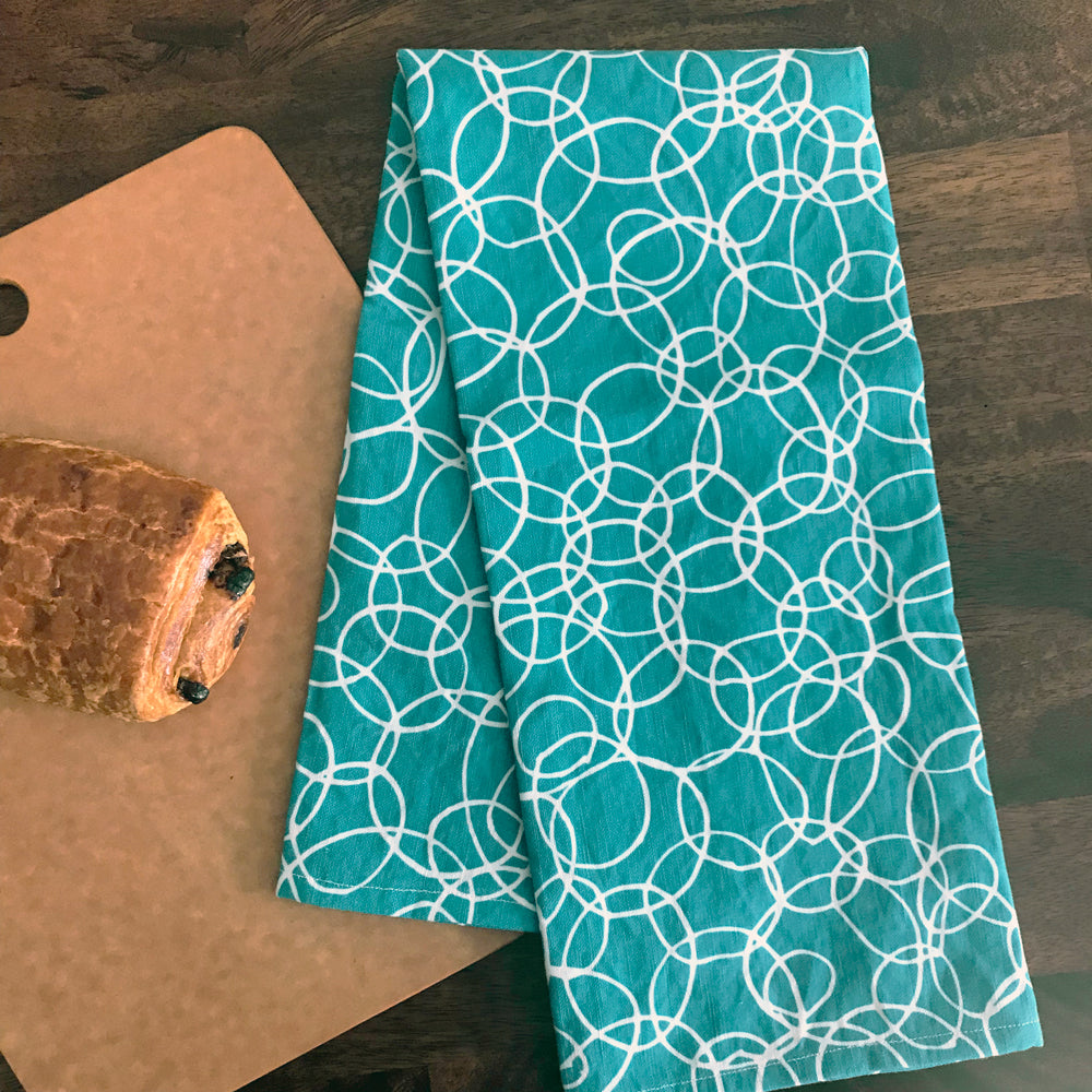 Turquoise and white scribble printed fun tea towel by Sunny Day Designs. Our "Bountiful Bubbles" printed dish towel is handmade in the USA from linen/cotton fabric. Shown folded in half on a wood table by a cutting board & chocolate croissant