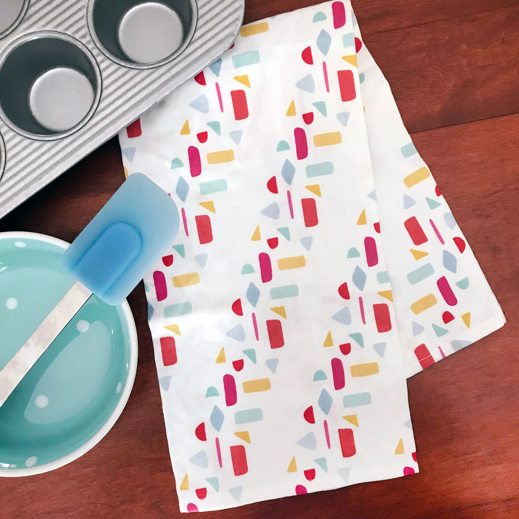 A colorfully printed geometric handmade kitchen tea towel is shown folded on top of a wooden table next to a muffin pan, a mixing bowl & a spatula. This fun dish towel makes a great bar towel!
