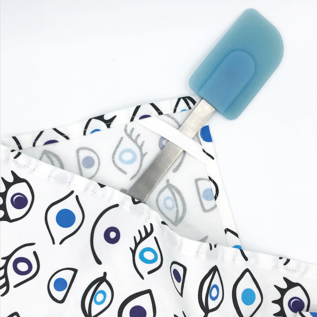 A handmade, linen/cotton fabric kitchen tea towel printed with a blue Evil Eye design is shown folded in half with a blue and silver spatula on a white background