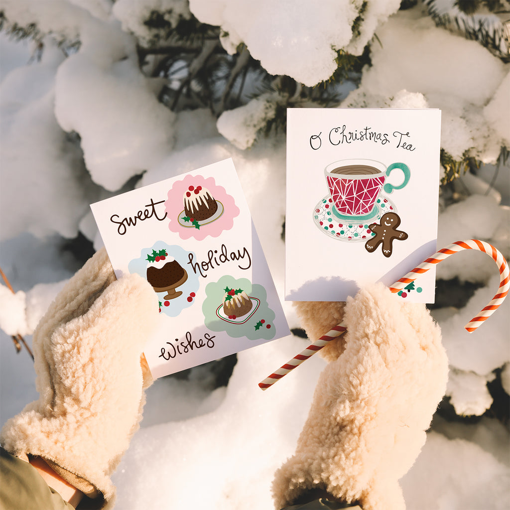 A pair of hands wearing fluffy ivory fleece mittens holds a candy cane & 2 Christmas cards by Sunny Day Designs outside in front of a snowy tree. The greeting cards held in their left hand is the Christmas Pudding Greeting Card, featuring 2 whimsical figgy pudding illustrations & hand lettered "Sweet Holiday Wishes" text. In the right hand they hold an O Christmas Tea card, featuring a holiday pun, an illustrated red/green/white patterned teacup, a gingerbread cookie & "O Christmas Tea" hand lettered text.