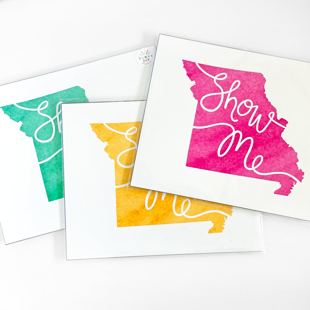 Set of three 'Show Me' Missouri art prints by Sunny Day Designs in vibrant watercolor styles. Each print features the outline of Missouri in different colors – teal, yellow, and pink – with the words 'Show Me' elegantly scripted across. The prints are displayed in protective packaging on a white background.