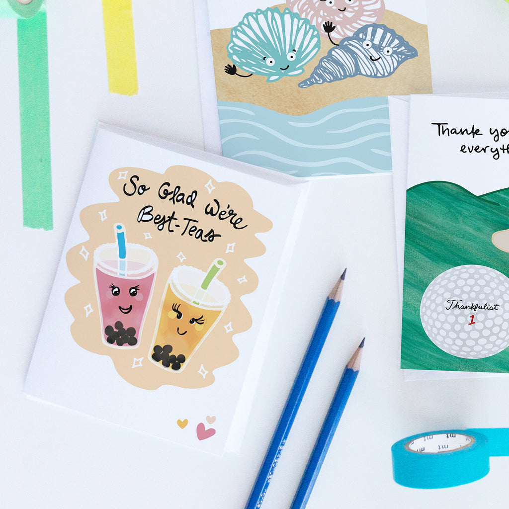 Colorful bubble tea-themed greeting card shown on a white table with colorful binder clips and a pencil. The greeting card is the "Best-Teas" Friendship greeting card by Sunny Day Designs, which is printed on sustainably sourced paper and features 2 colorful, smiling bubble tea drinks on a tan and white background with 3 hearts in the bottom right corner. The hand lettered  text on the front of the card reads "So Glad We're Best-Teas"