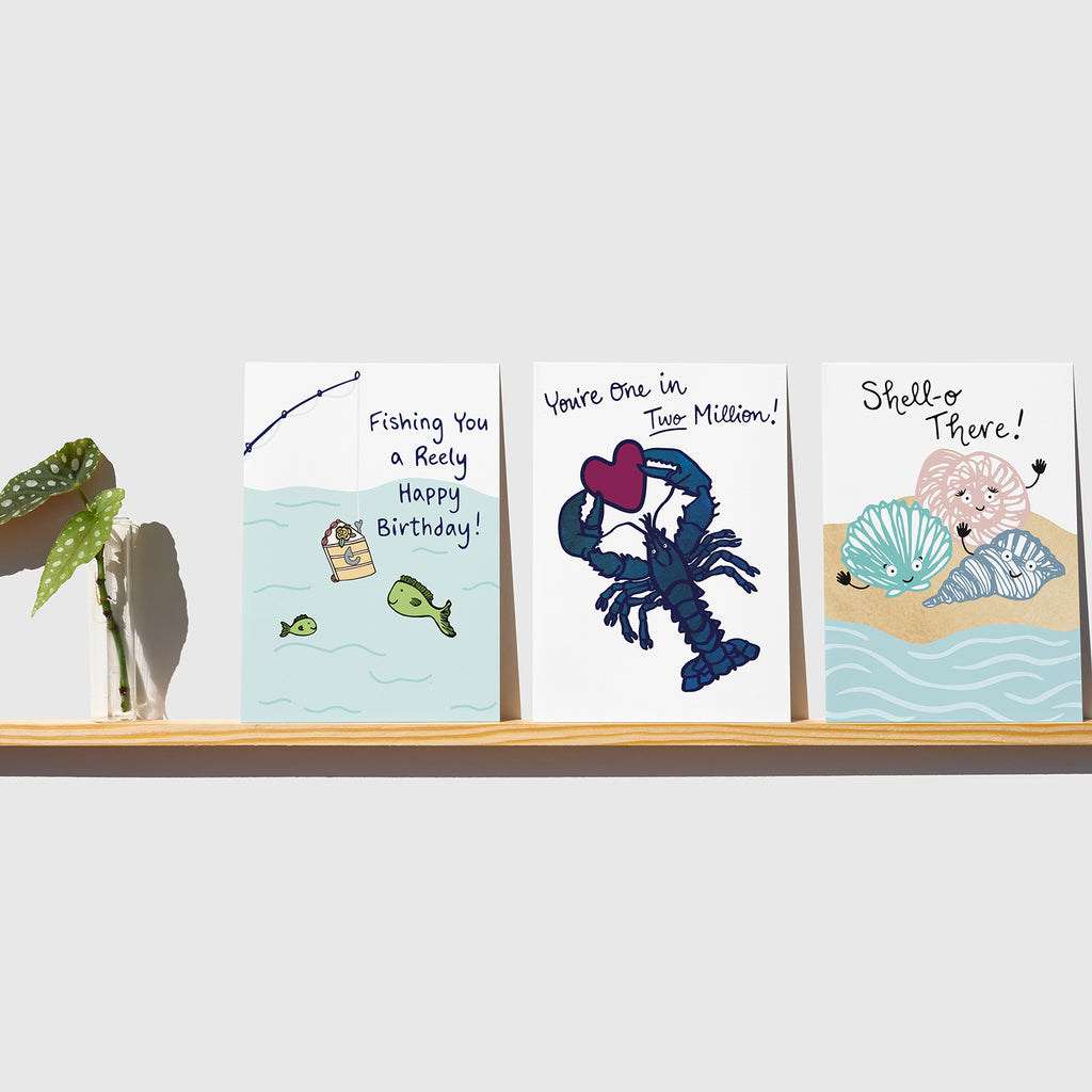 A wooden shelf attached to a white wall holds 3 colorful coastal greeting cards by Sunny Day Designs next to a small vase with a green leafy plant. From left to right, the greeting cards displayed are the Fishing You A Reely Happy Birthday card (a pun card featuring a fishing pole dangling a slice of birthday cake into the water while 2 fish swim nearby), the One In Two Million Blue Lobster greeting card (featuring a rare blue lobster holding a magenta purple heart) & the Shell-o There! seashell pun card.