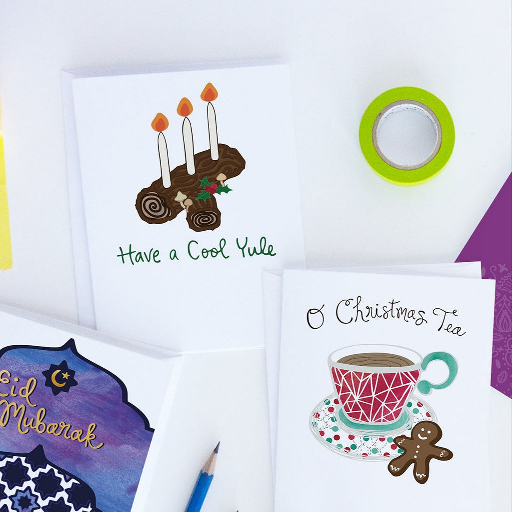 3 colorful holiday greeting cards by Sunny Day Designs are shown on a white background next to a blue pencil and a roll of green washi tape. The holiday cards shown are: a purple, blue & gold watercolor sunset-themed Eid Mubarak greeting card for Eid al-Fitr/Ramadan, a Have A Cool Yule Winter Solstice Pagan greeting card featuring a Yule Log and 3 candles, and an O Christmas Tea holiday pun Christmas card featuring a red, green, and white illustration of a patterned tea cup and a gingerbread man cookie.