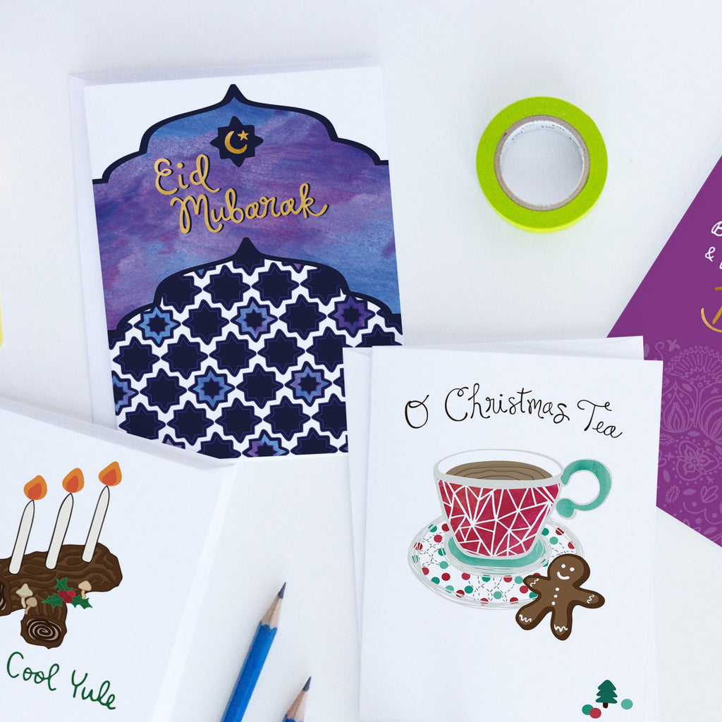 4  holiday greeting cards by Sunny Day Designs are shown laying on a white background with pencils and a roll of green washi tape. The cards shown are the Eid Mubarak watercolor sunset-inspired greeting card for Eid al-Fitr/Ramadan, the Have A Cool Yule Pagan greeting card for the Winter Solstice, the O Christmas Tea food-inspired Christmas card showing a cup of tea and a gingerbread man cookie, and our purple and gold Bright & Beautiful Diwali greeting card. All 4 cards are made in the USA.