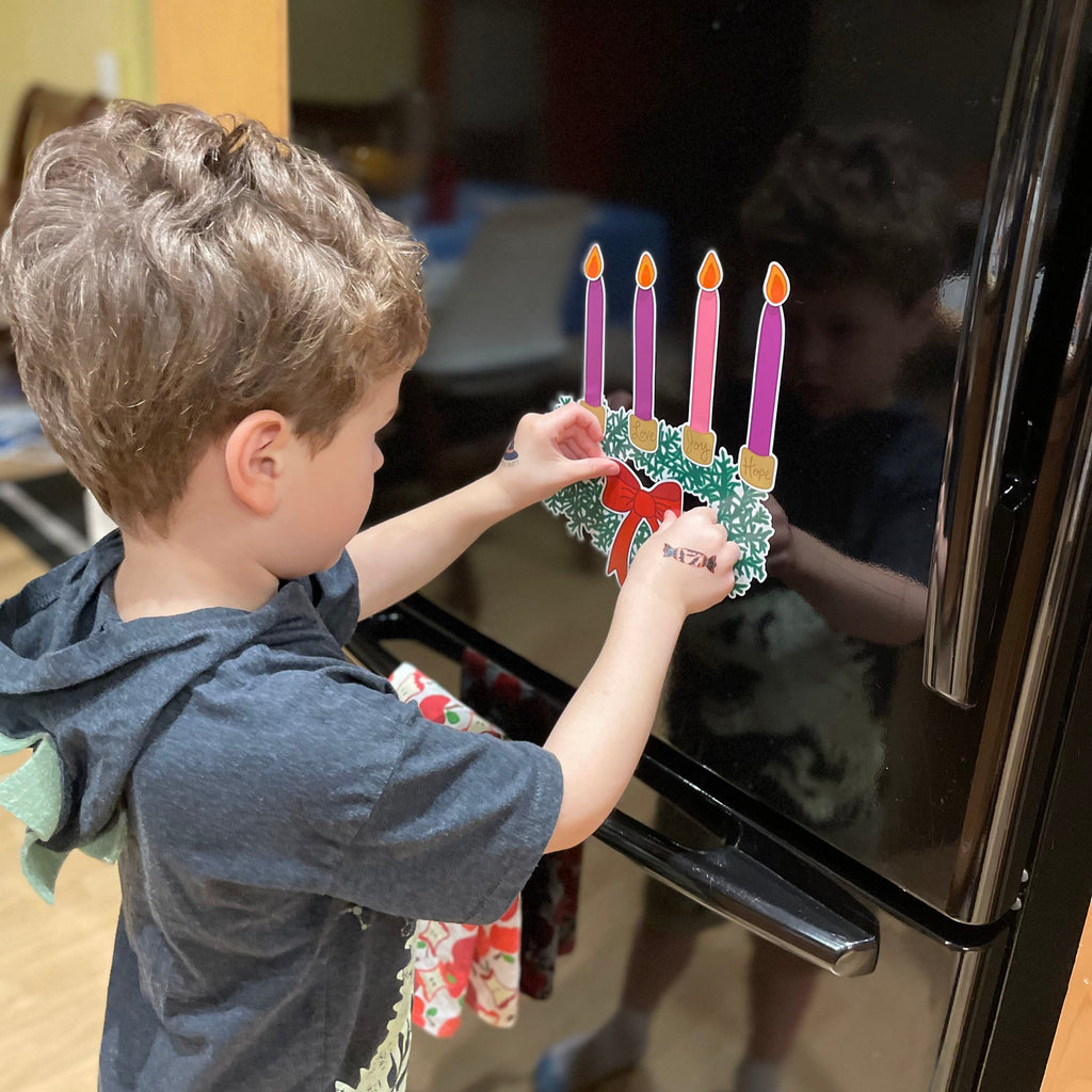 A young boy child safely uses a flame-free, reusable Advent Wreath holiday countdown vinyl magnet set by Sunny Day Designs on the black refrigerator in his family's kitchen as he places purple and pink magnetic candles and a red bow magnet onto a green magnetic Advent Wreath to help him count down the days leading up to Christmas during the Christian season of Advent. This fun holiday magnet set is also made in the USA.