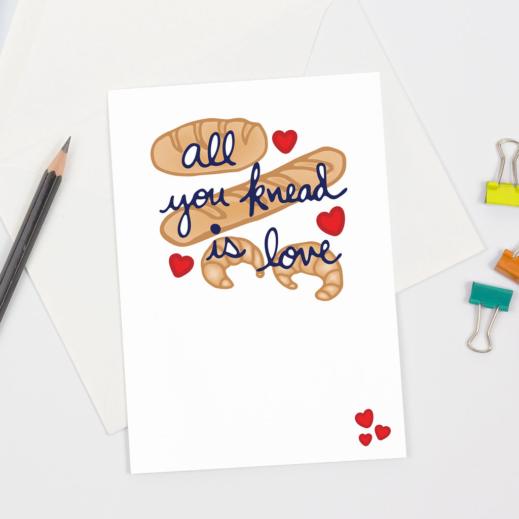 Minimalist and playful greeting card featuring hand-drawn croissants and baguettes with navy blue cursive text that reads “All you knead is love,” accented by red hearts. Clean white background with flat lay composition, styled alongside a pencil, envelope, and colorful paper clips  perfect for foodies, bakers, or pun lovers.