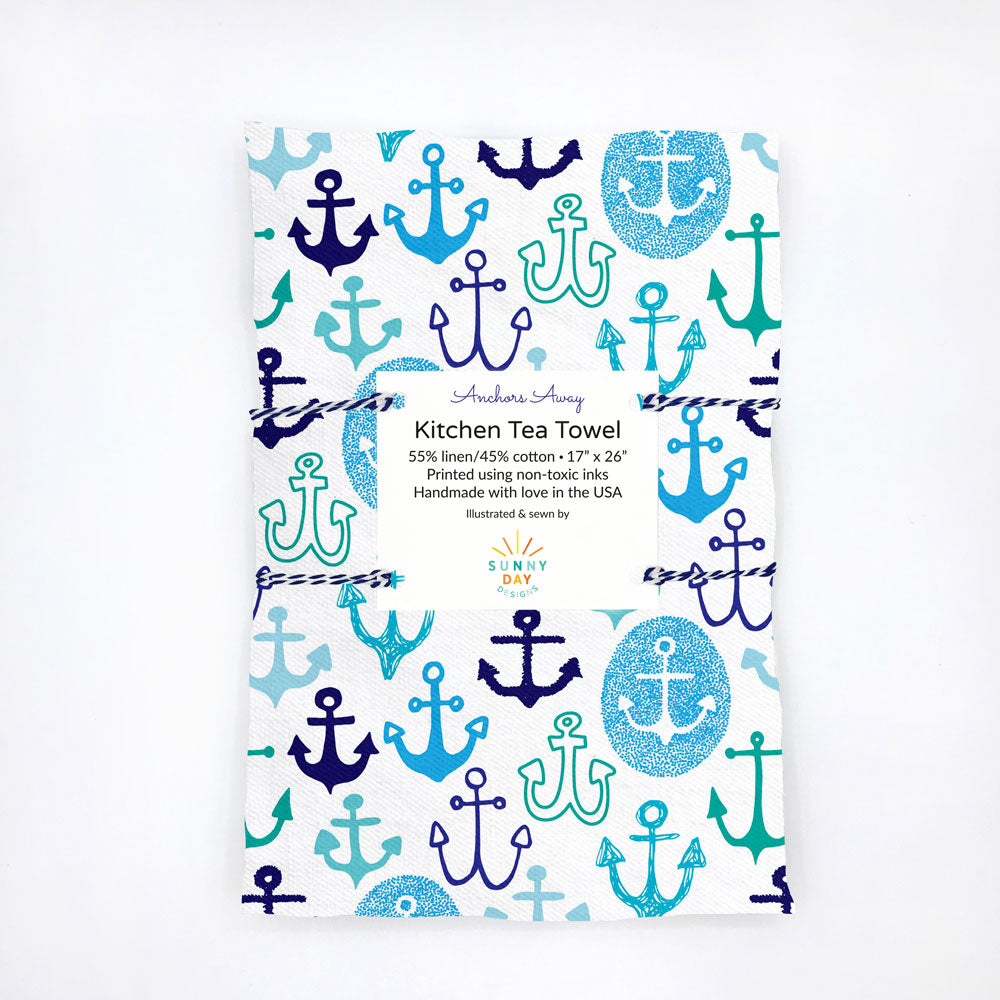 Packaged 'Anchors Away' kitchen tea towel by Sunny Day Designs, featuring a nautical anchor pattern in various shades of blue and teal on a white background. The towel is made from a 55% linen and 45% cotton blend, printed with non-toxic inks, and is handcrafted in the USA.