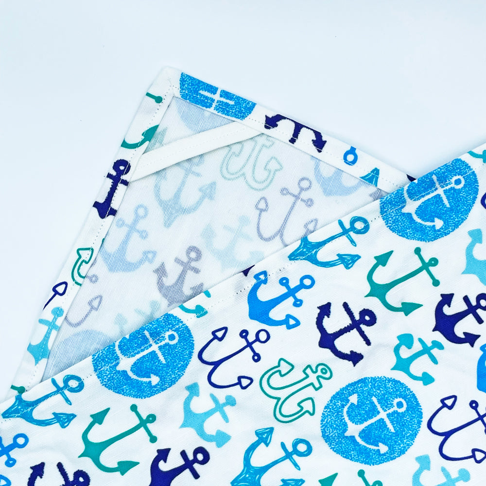 Close-up of the 'Anchors Away' kitchen tea towel from Sunny Day Designs, featuring a vibrant pattern of anchors in shades of blue, teal, and navy on a white background. The towel includes a hanging loop stitched into the corner for convenient storage. Made from a linen and cotton blend.