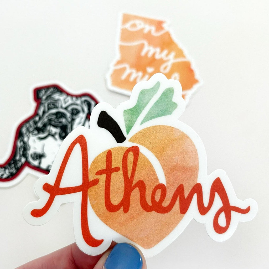 An orange, peach, green and brown Athens, Georgia peach-themed UGA die cut vinyl sticker by Sunny Day Designs is shown held in a hand in front of a white background, with 2 other University of Georgia and Georgia themed stickers shown in the far background.