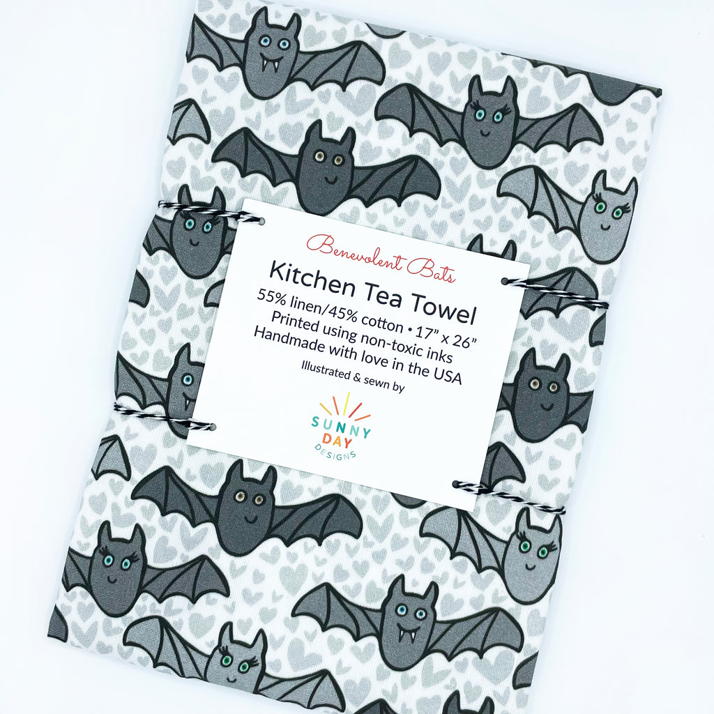 A tilted view of our cute, handmade, Halloween bat-printed linen/cotton kitchen tea towel - this photo shows a variety of playful, Benevolent Bats in shades of gray with light gray hearts in the background.