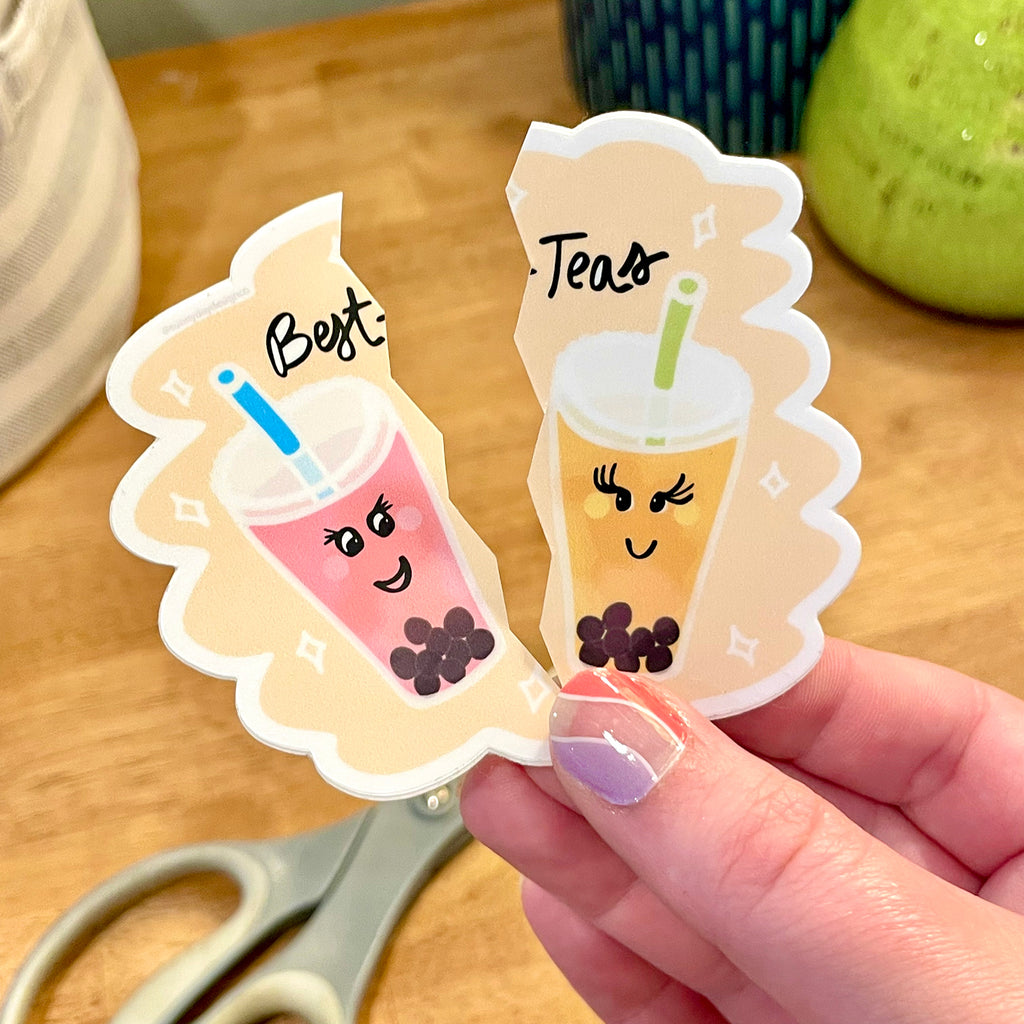 A hand holding a Best-Teas Vinyl Sticker by Sunny Day Designs that has been cut in half (to create a BFF sticker where each half of the sticker goes to one of the best friends). A sticker is also shown in the background.