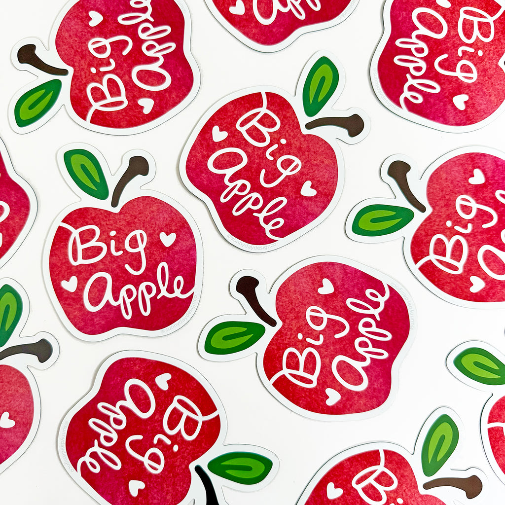 A group of 12 apple-shaped NYC inspired Big Apple souvenir magnets are shown laying scattered across a white background. These fun New York City magnet souvenirs make great small gifts. They're designed by Sunny Day Designs and are made in the USA in small batches.