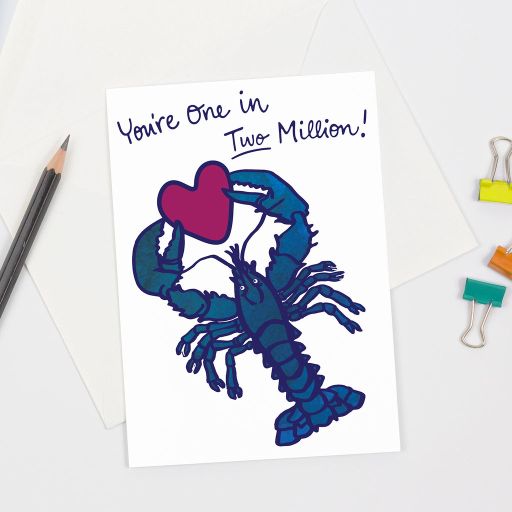 Greeting card with a blue water color lobster holding a magenta heart, with the text 'You're One in Two Million!' displayed above. The card is shown with a white envelope, pencil, and colorful binder clips arranged around it.