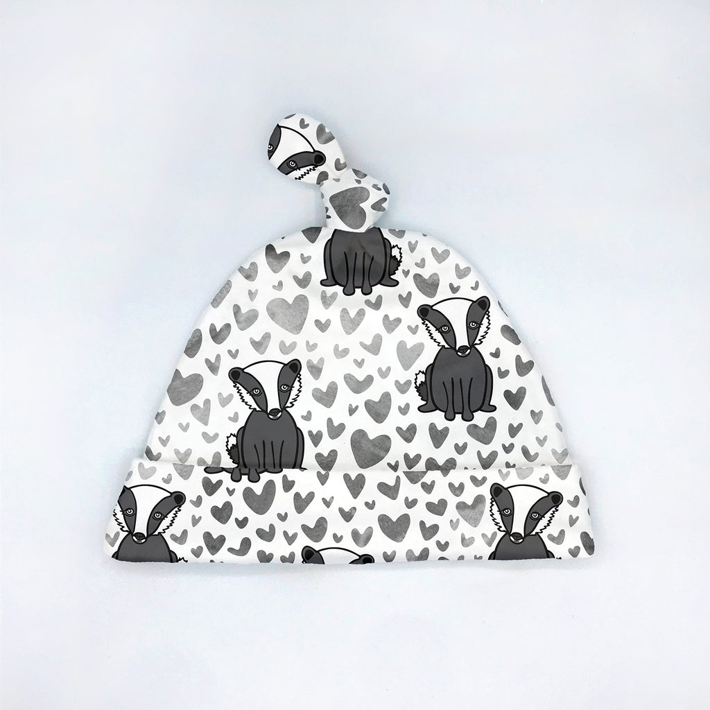 A white baby hat with a tie top "top knot" design featuring a repeating animal-themed pattern of black and white badgers surrounded by watercolor hearts in shades of grey. This adorable badger printed baby hat will appeal to animal lovers as well as Wisconsinites and University of Wisconsin fans! Displayed flat on a clean white background, this cute organic newborn hat is part of the Bonnie Badgers eco-friendly baby apparel collection by Sunny Day Designs.