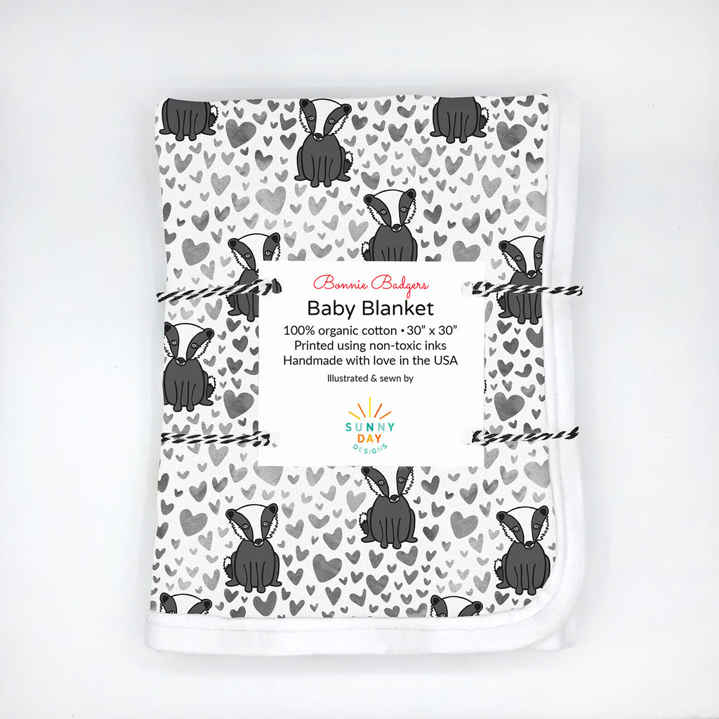 A folded organic cotton baby blanket featuring a black and white badger animal  print design with scattered grey watercolor heart is shown packaged with a white card label that reads “Bonnie Badgers Baby Blanket – 100% organic cotton, 30” x 30”, printed using non-toxic inks, handmade in the USA.” This cute Wisconsin badger baby blanket by Sunny Day Designs makes a perfect baby shower gift for University of Wisconsin Madison alumni, students & fans.