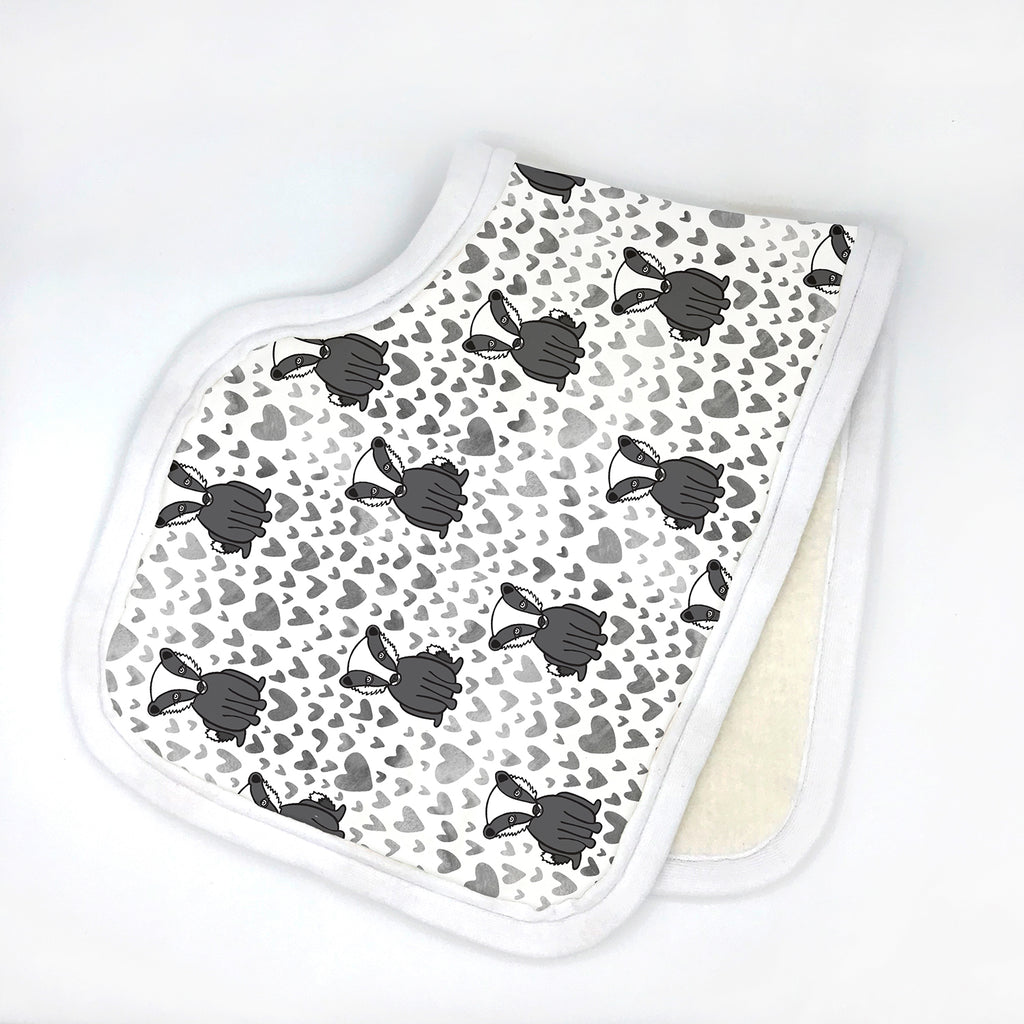 Contoured white baby burp cloth featuring a repeating pattern of black and white Bonnie Badgers with scattered grey hearts. Displayed folded in half on a white background with soft white trim, part of the Bonnie Badgers eco-friendly collection by Sunny Day Designs.