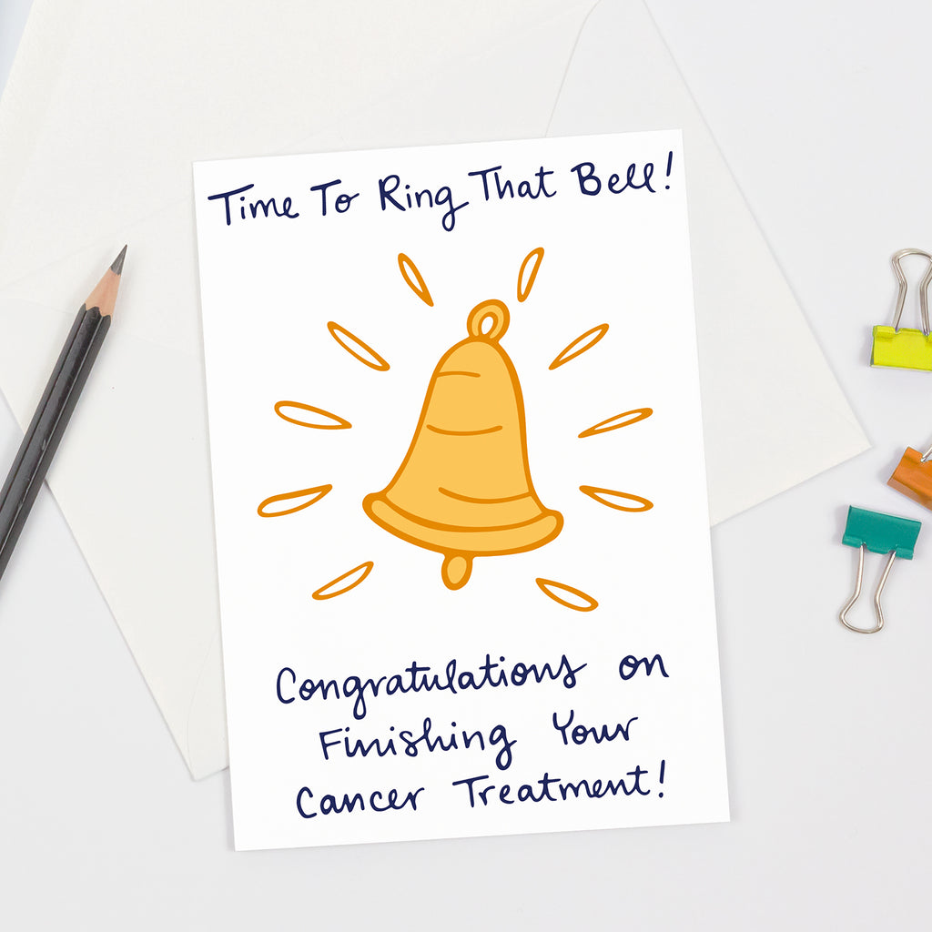 Greeting card featuring a hand-drawn yellow bell with radiating lines and the message “Time To Ring That Bell! Congratulations on Finishing Your Cancer Treatment!” in playful handwritten-style font. This card is perfect to send a congratulations message to a cancer patient who just finished the end of their cancer treatment! Displayed with a pencil, envelope, and colorful paper clips on a clean white background.