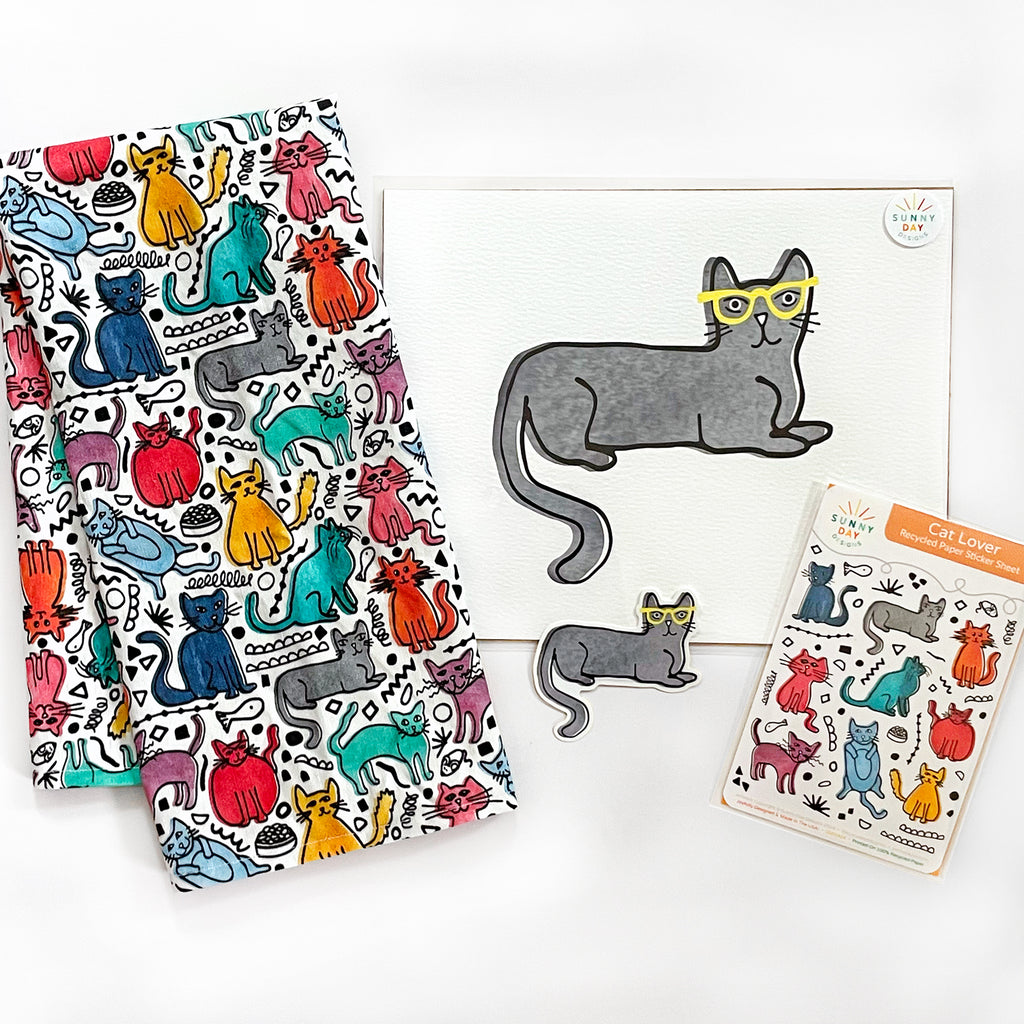 4 colorful, cat-themed gifts are shown on a white background - a gift set that any cat lover would love, which includes a Friendly Felines colorful tea towel, an art print of a gray cat wearing yellow glasses, a vinyl sticker of a gray cat wearing yellow glasses, and a recycled paper sticker sheet featuring colorful cats. All products designed by Sunny Day Designs.