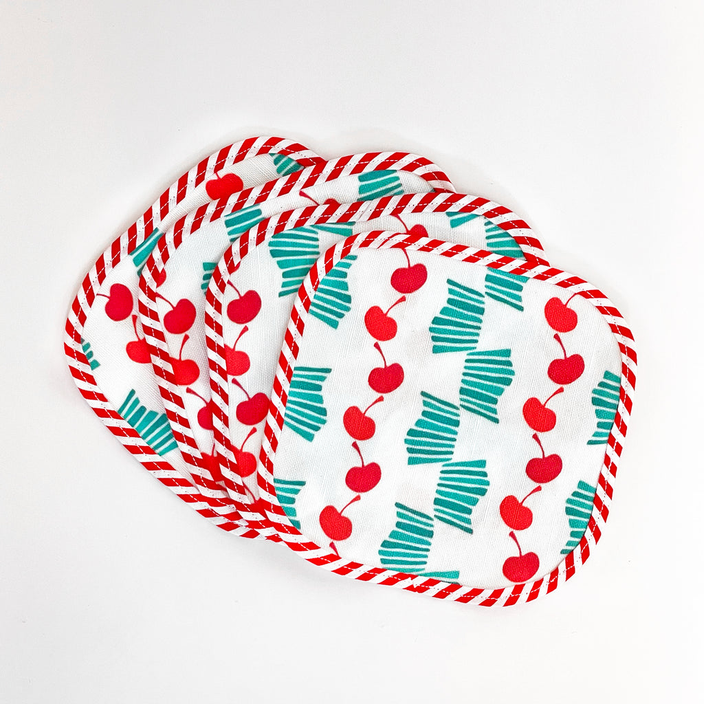 Fanned-out set of four Cheerful Cherries fabric cocktail napkins. The napkins feature vibrant red cherry patterns and teal striped bows on a white background, edged with red and white striped trim, perfect for adding a fun touch to any table setting.
