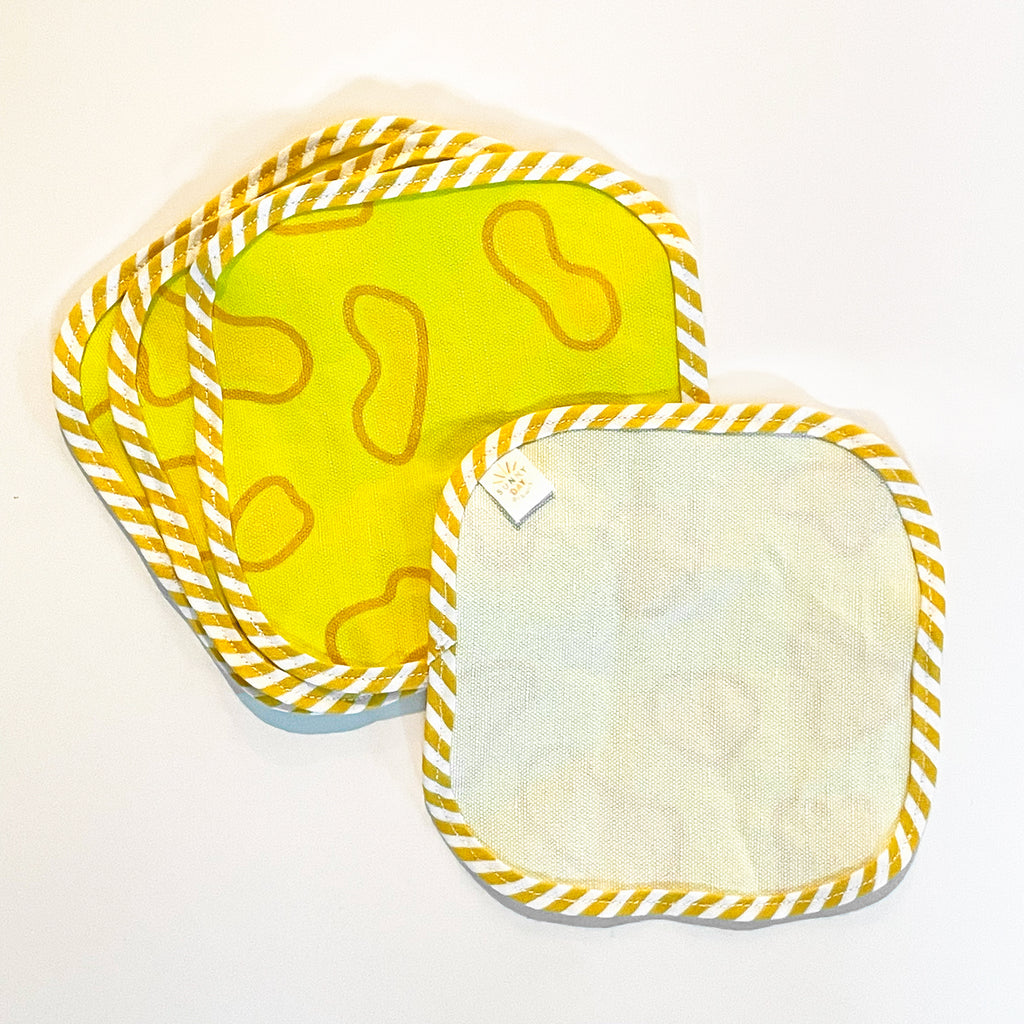 Set of four yellow fabric cocktail napkins with playful cheese-inspired patterns, fanned out. The napkins are bordered with yellow and white striped trim, adding a whimsical touch. One napkin is flipped to show the back side with a label.
