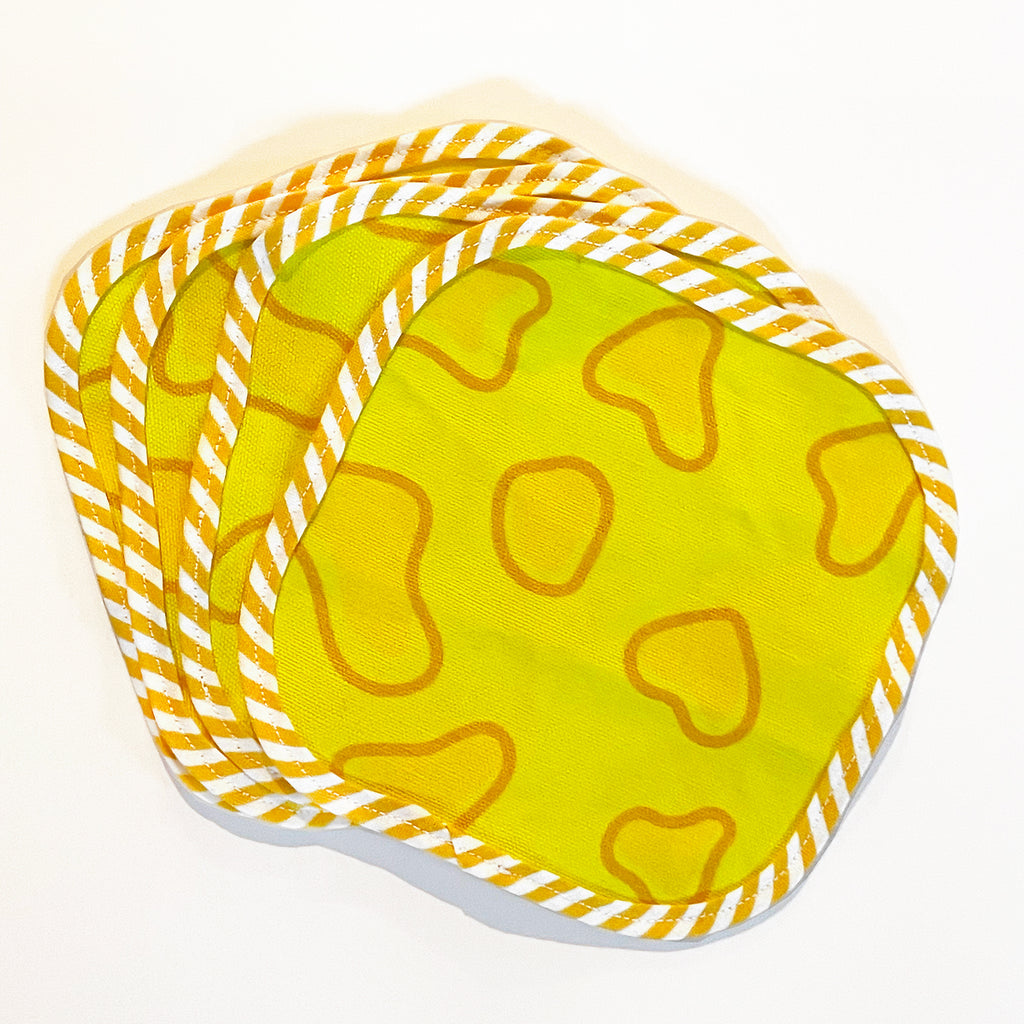 Four fabric cocktail napkins with a bright yellow background and cheese-inspired designs, fanned out. The edges are finished with a yellow and white striped trim, adding a bold and fun element to the set.