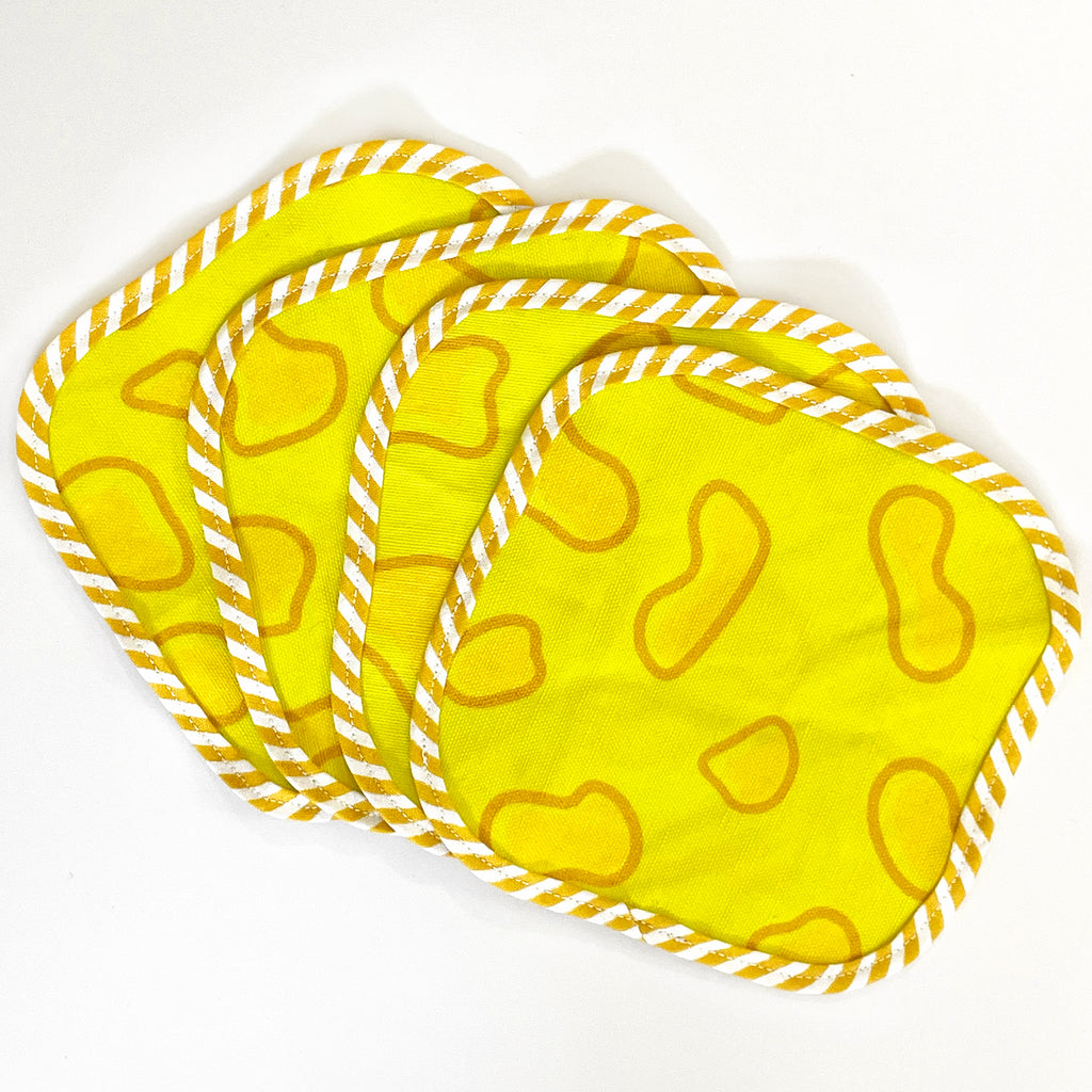 Four fabric cocktail napkins with a bright yellow background and cheese-inspired designs, fanned out. The edges are finished with a yellow and white striped trim, adding a bold and fun element to the set