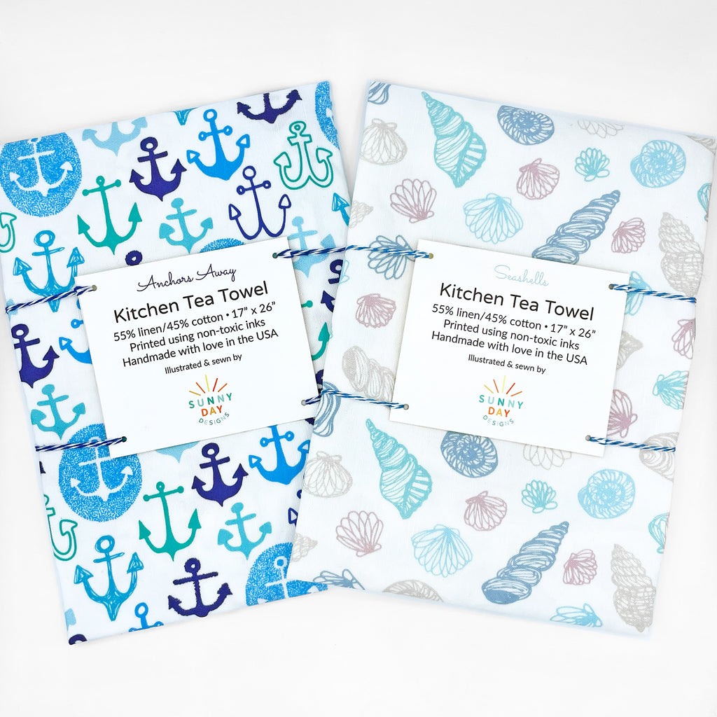 Set of two packaged kitchen tea towels from Sunny Day Designs, featuring coastal themes. One towel showcases a blue and teal 'Anchors Away' design with various anchor illustrations, while the other 'Seashells' design displays a pattern of pastel seashells in soft blue, teal, and pink shades. Both towels are made of 55% linen and 45% cotton, printed with non-toxic inks, and handmade in the USA.