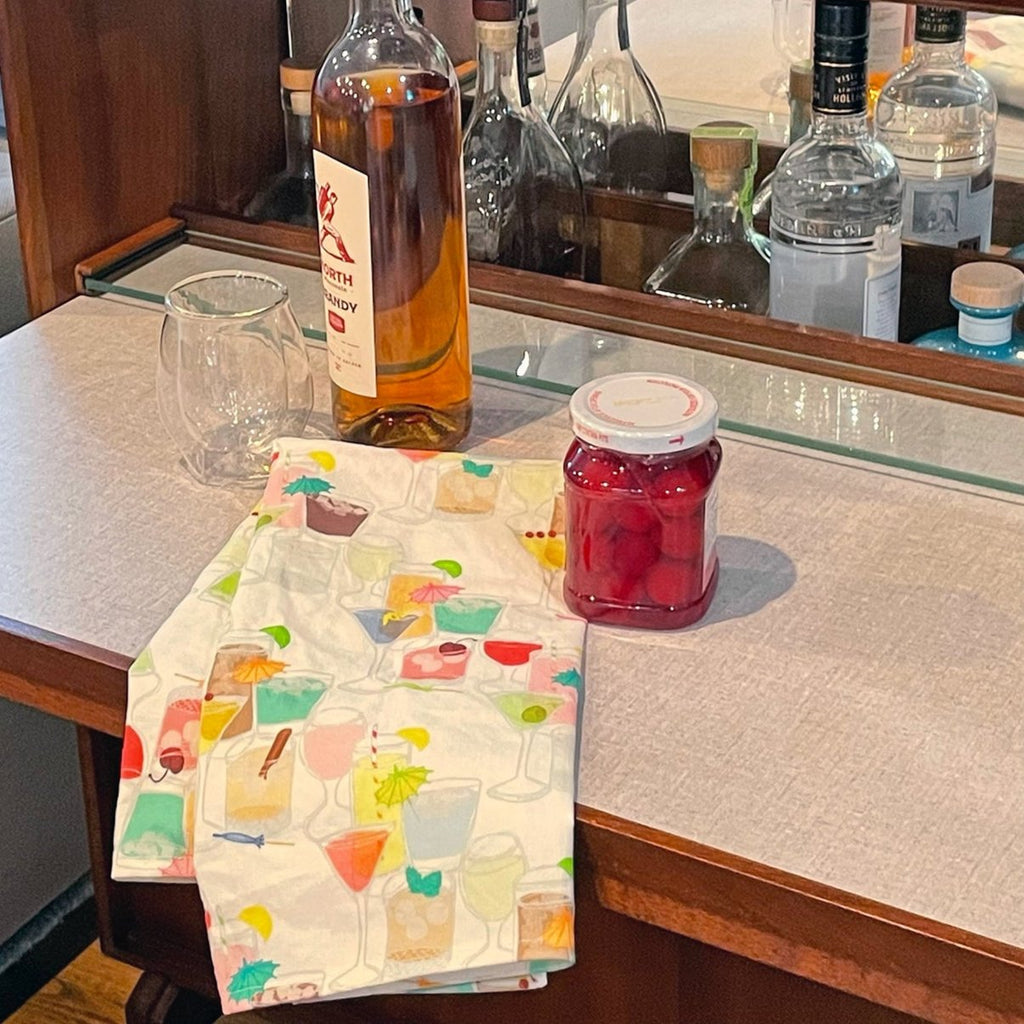 A colorful & cute retro-inspired cocktail & alcoholic drink-themed linen/cotton bar towel by Sunny Day Designs is shown draped on the counter of a vintage wooden home bar cabinet, next to several bottles of alcohol, a jar of cherries, and an empty old fashioned cocktail glass.