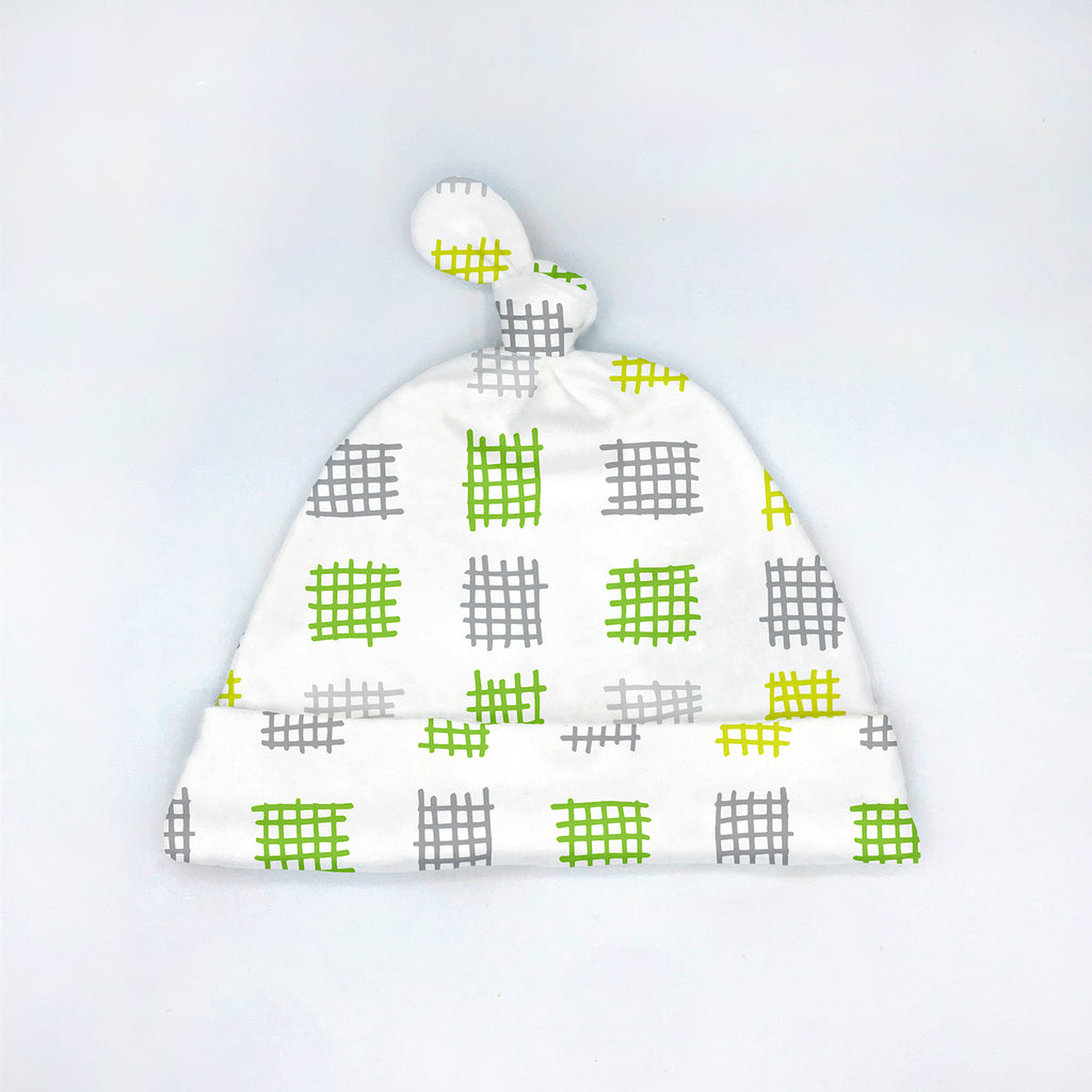 A handmade tie-top newborn beanie-style baby hat with a green, gray and white abstract checkered geometric print design titled "Criss-Cross Applesauce" is shown laying flat on top of a white background.. This soft 0-3M sized baby hat is gender neutral and joyfully designed and made in the USA by Sunny Day Designs.