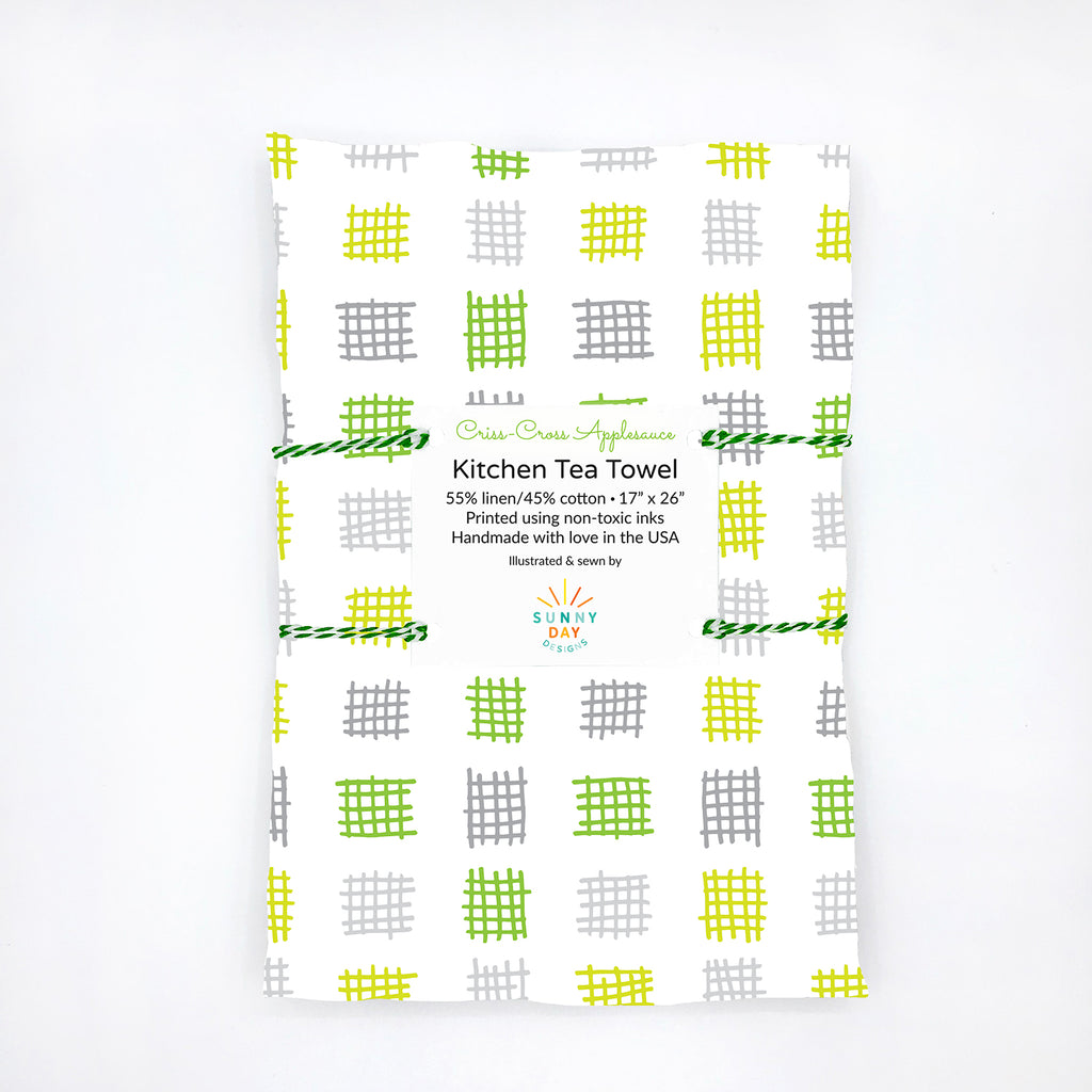 Folded kitchen tea towel featuring a criss-cross grid pattern in lime green, yellow, and grey on a white background, made from 55% linen and 45% cotton. Packaged with a white label that reads “Criss-Cross Applesauce Kitchen Tea Towel – 55% linen / 45% cotton, 17” x 26”, printed using non-toxic inks, handmade with love in the USA.” Minimalist flat lay on a white background with eco-friendly branding by Sunny Day Designs.