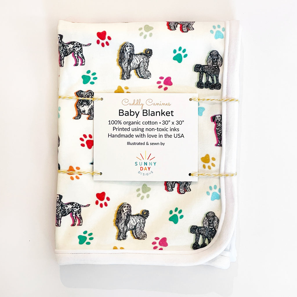 A 30x30-inch organic cotton baby blanket featuring illustrations of various dog breeds and colorful paw prints, packaged with a label from Sunny Day Designs. The print includes 7 adorable dog breeds sketched in black on a white fabric background and accented with colorful paw prints and highlights in yellow, orange, green, turquoise & light blue.