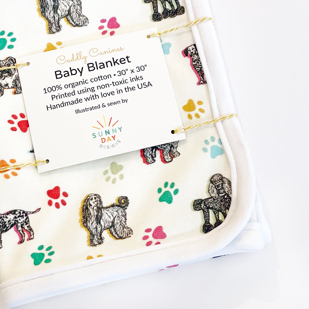 A close-up tilted view of the baby blanket with dog illustrations, highlighting the Sunny Day Designs product label and colorful lettering design. The blanket print includes 7 adorable dog breeds sketched in black on a white fabric background and accented with colorful paw prints and highlights in yellow, orange, green, turquoise & light blue.