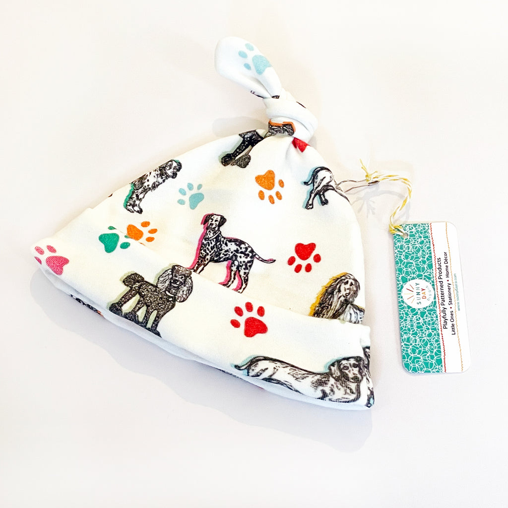A baby hat featuring dog illustrations and colorful paw prints. The print  includes 7 adorable dog breeds sketched in black on a white fabric background and accented with colorful paw prints and highlights in yellow, orange, green, turquoise & light blue. Displayed with a tag attached by string, from Sunny Day Designs.