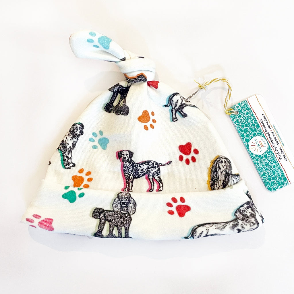 Cuddly Canines dog printed organic cotton baby hat by Sunny Day Designs features 7 cute dog breeds on a white background with colorful paw prints. The print  includes 7 adorable dog breeds sketched in black on a white fabric background and accented with colorful paw prints and highlights in yellow, orange, green, turquoise & light blue. with a tag attached by string, from Sunny Day Designs.