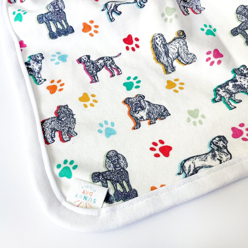 A detailed corner shot of the baby blanket, focusing on the dog breed illustrations and multicolored paw prints on a white background.The print  includes 7 adorable dog breeds sketched in black on a white fabric background and accented with colorful paw prints and highlights in yellow, orange, green, turquoise & light blue. corner includes mini fabric "Sunny Day Designs" tag.